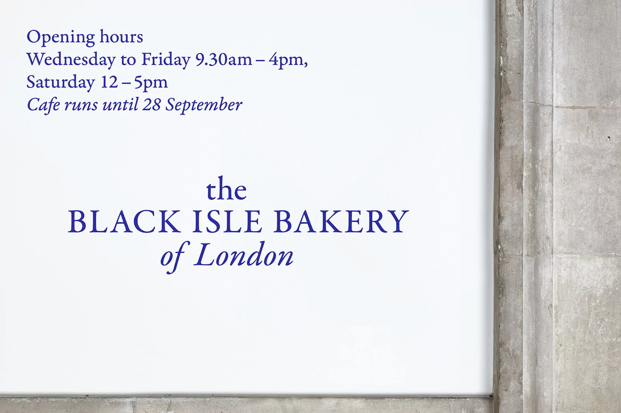The Black Isle Bakery of London sign on a white wall with opening hours in blue text, next to a grey concrete wall.