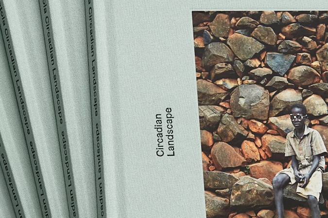Closed hardcover book titled Circadian Landscape next to a photograph of a boy seated against a stacked stone wall.