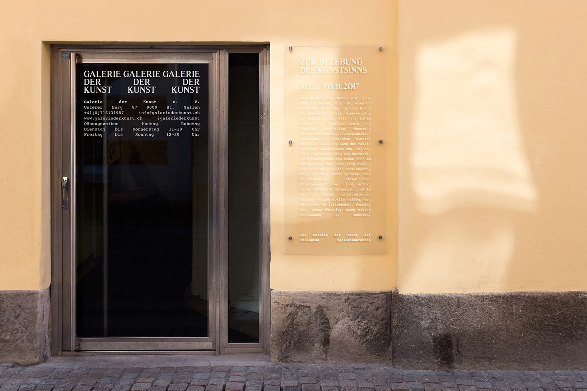 Exterior view of Galerie Der Kunst featuring the gallery's entrance door, a promotional sign, and the cobblestone pavement.
