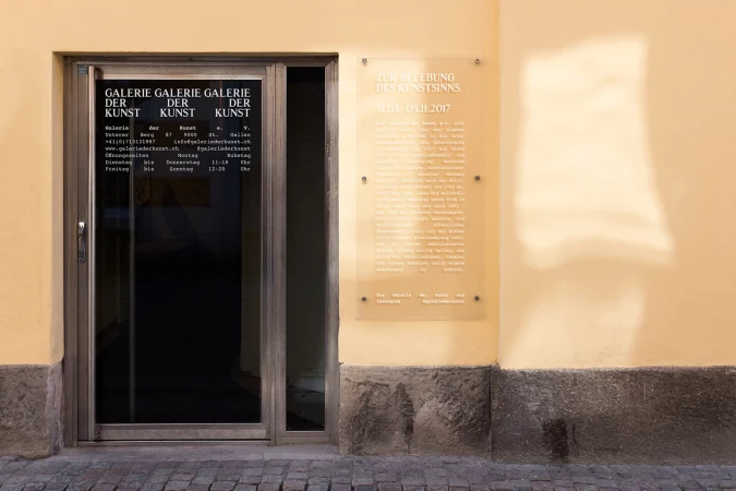 Exterior view of Galerie Der Kunst featuring the gallery's entrance door, a promotional sign, and the cobblestone pavement.
