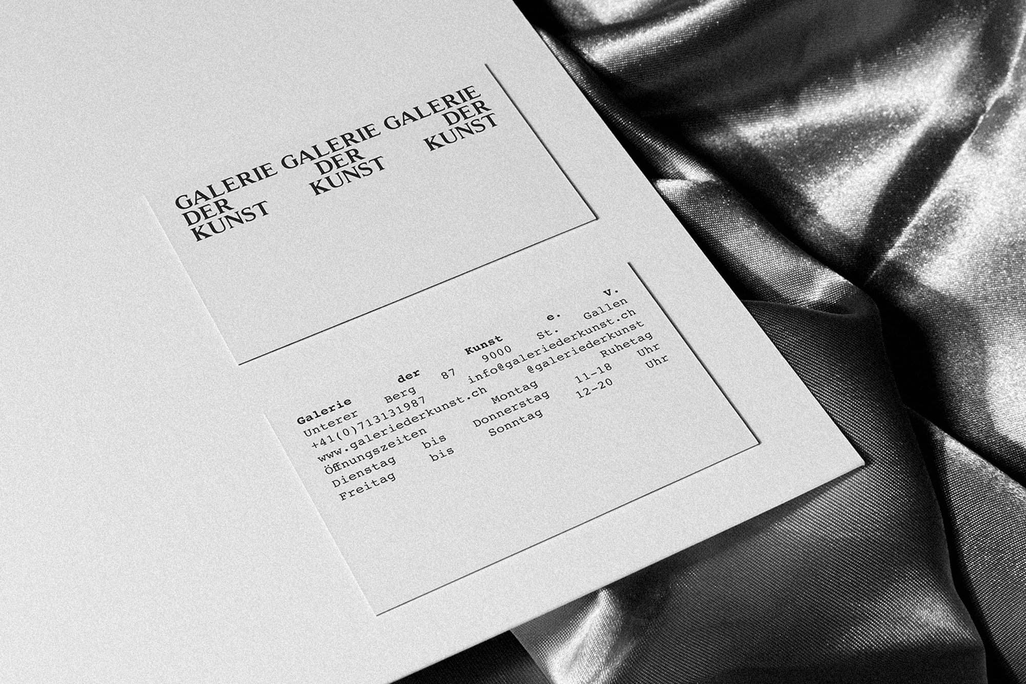 Close-up of a flyer for Galerie der Kunst art gallery featuring its name, address, and contact details. Shot on a silver satin background.