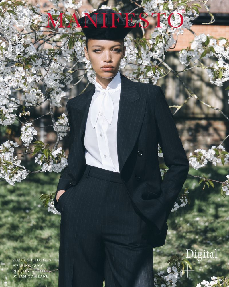 A woman poses outdoors wearing a dark pin-stripe suit, black beret, and white blouse, standing in front of a blooming white flowering tree.
