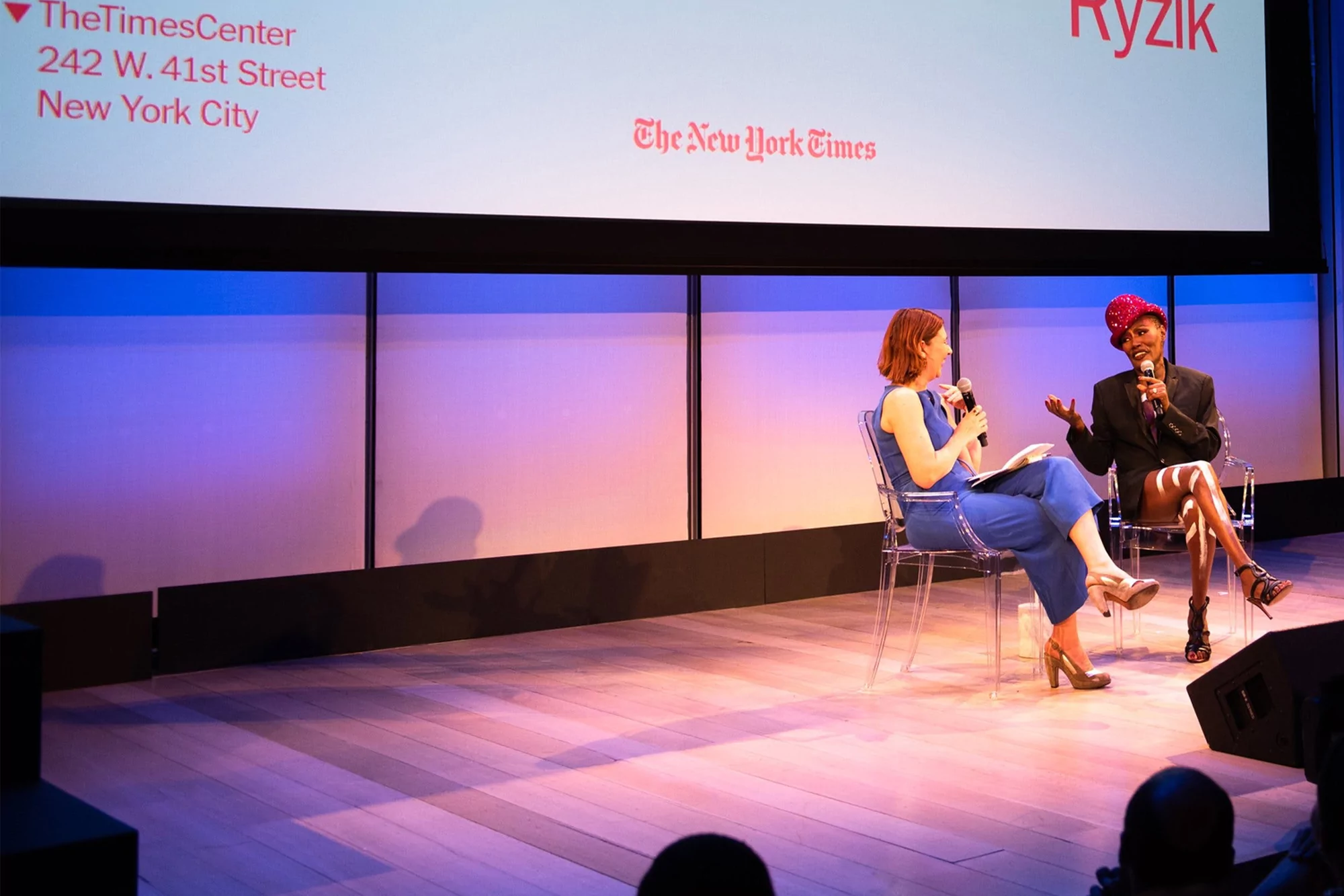 Two women on stage during a New York Times event; one interviewing the other, both holding microphones in a lit auditorium.