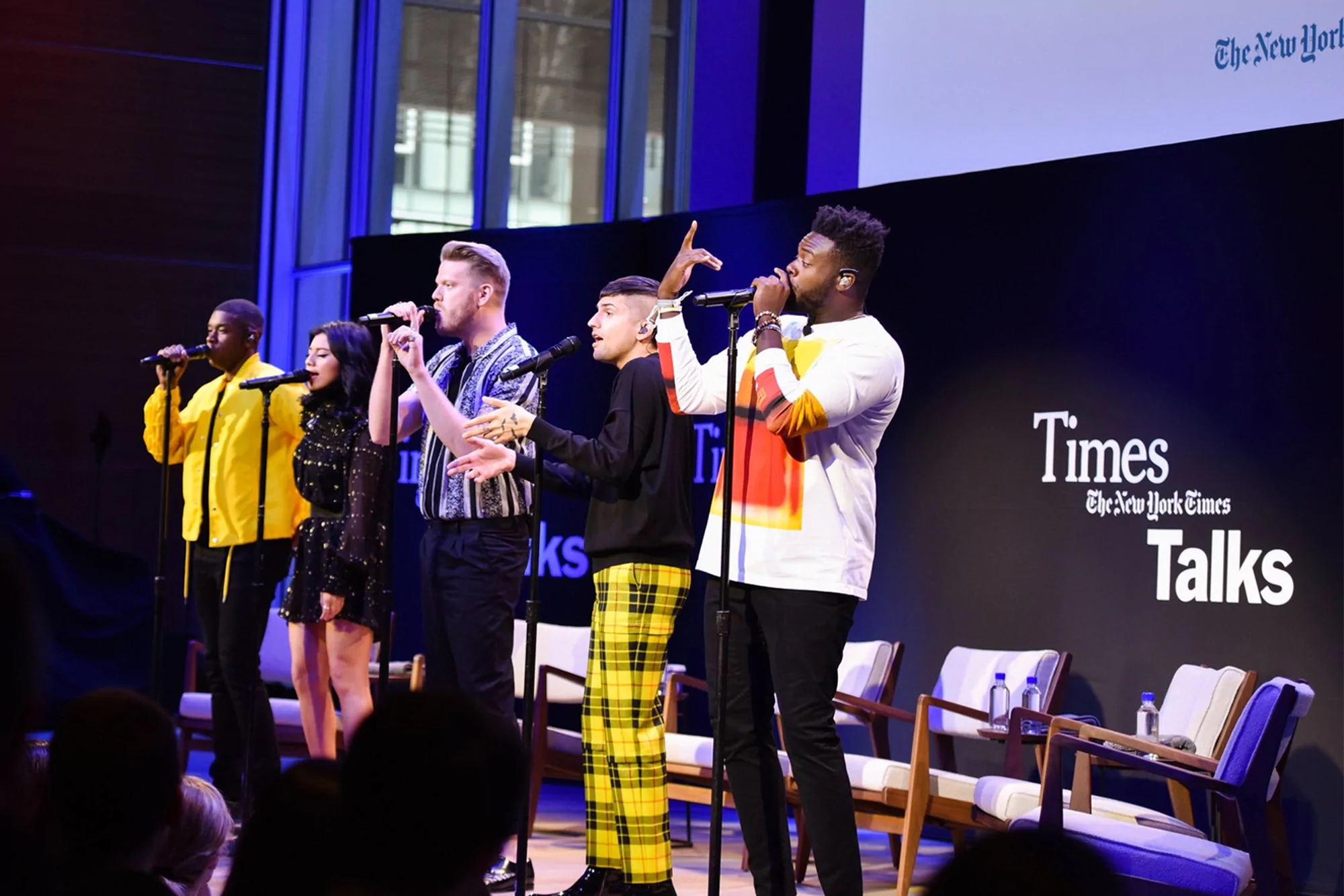 Five vocalists performing on stage at The New York Times Talks event; the diverse group sings into microphones on a stage with chairs.