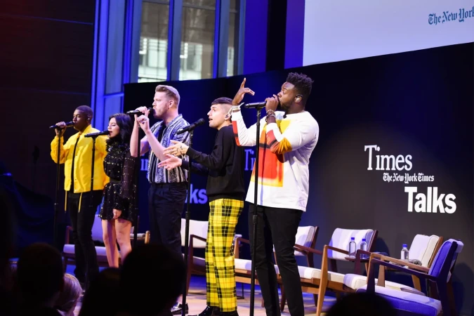 Five vocalists performing on stage at The New York Times Talks event; the diverse group sings into microphones on a stage with chairs.