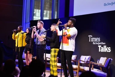 Five performers singing into microphones on stage during a TimesTalks event with branding visible in the background