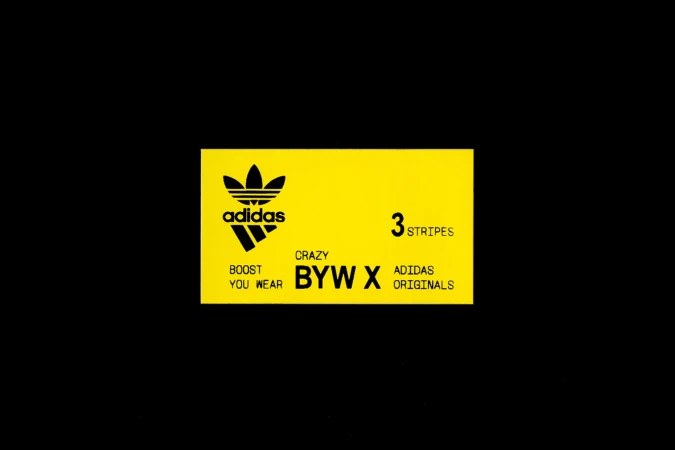Adidas brand logo and text on a yellow rectangle against black background. Features the Adidas trefoil and '3 stripes' tagline.