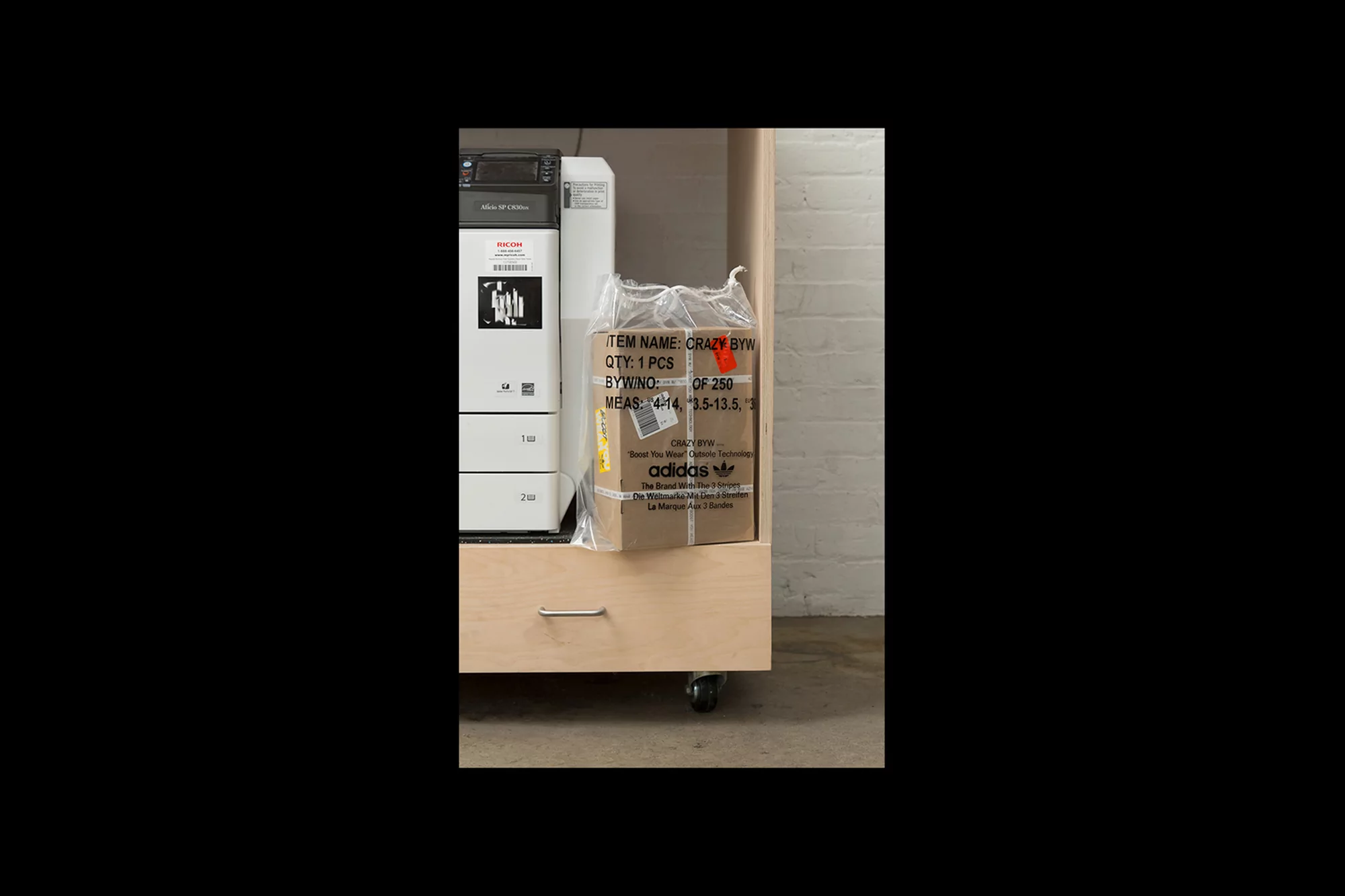 Adidas shoe box in plastic bag inside a light wood cabinet next to a white Ricoh printer with gray concrete floor and wall.