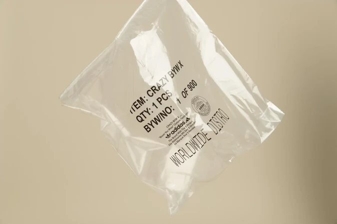 Clear plastic bag with Adidas logo and product information, neutral background. Packaging features black printed text and branding.