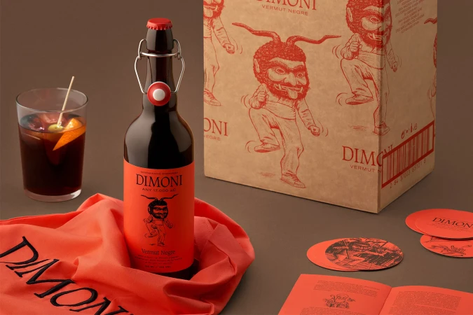 Bottle of Dimoni Vermut Negre beside a glass of vermouth with an orange slice and olive, box, book, and coasters on a table.