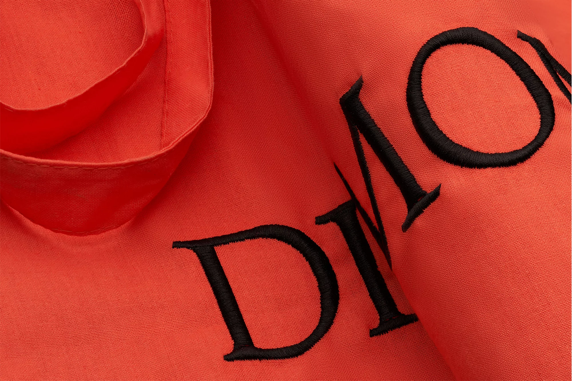 Close-up of an orange tote bag with black embroidered lettering. The fabric is folded and textured, creating a textured, vibrant graphic.