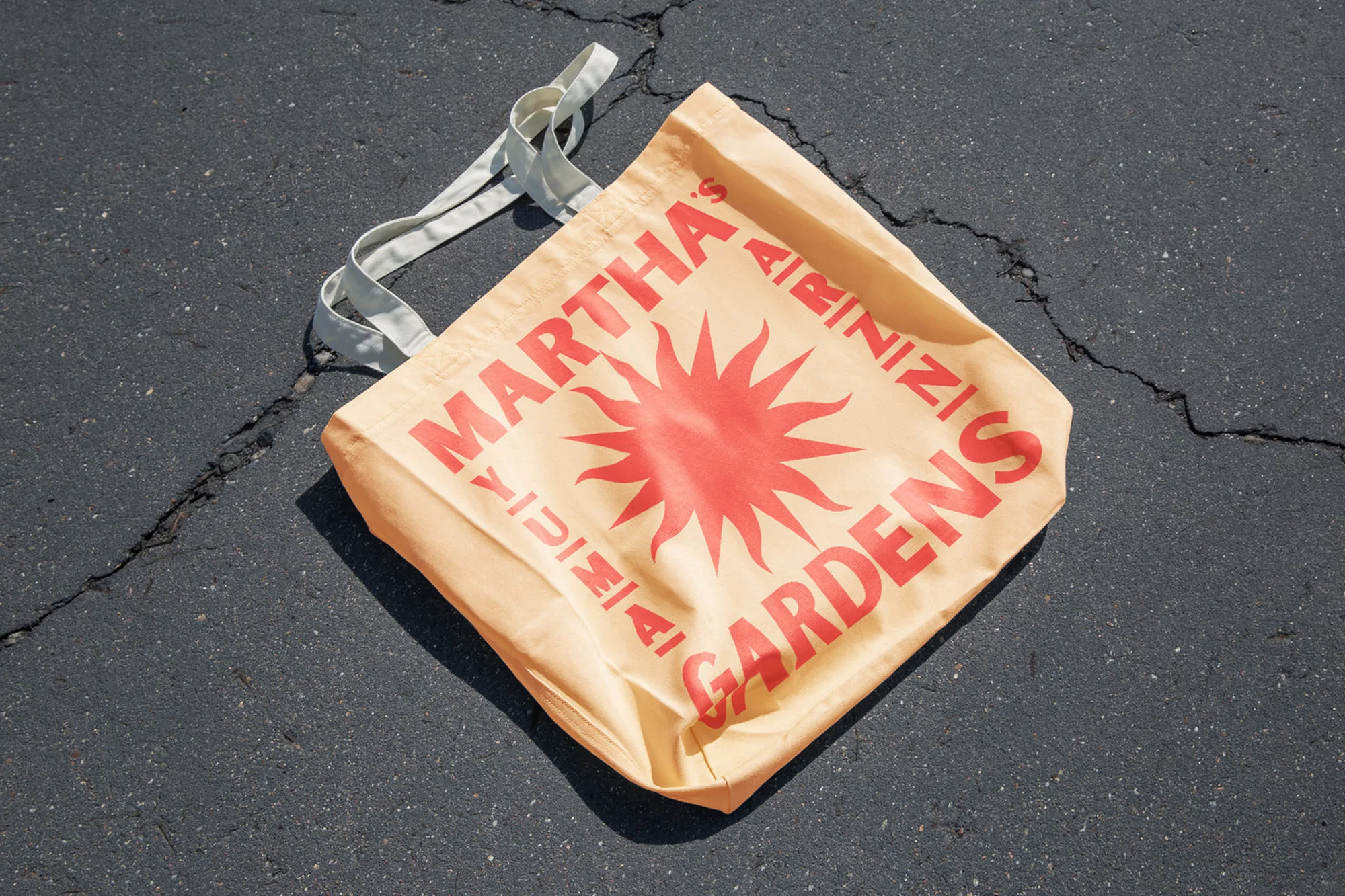 Canvas tote bag with red 'Martha's Gardens' text and sun design on asphalt background, showcasing style and utility.