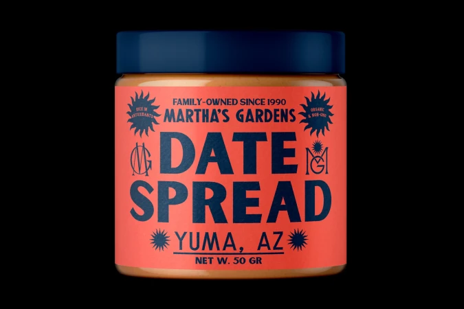 Eye-level studio shot of a jar of Martha's Gardens Date Spread, with a coral-colored label and dark blue lid, against a stark black backdrop.