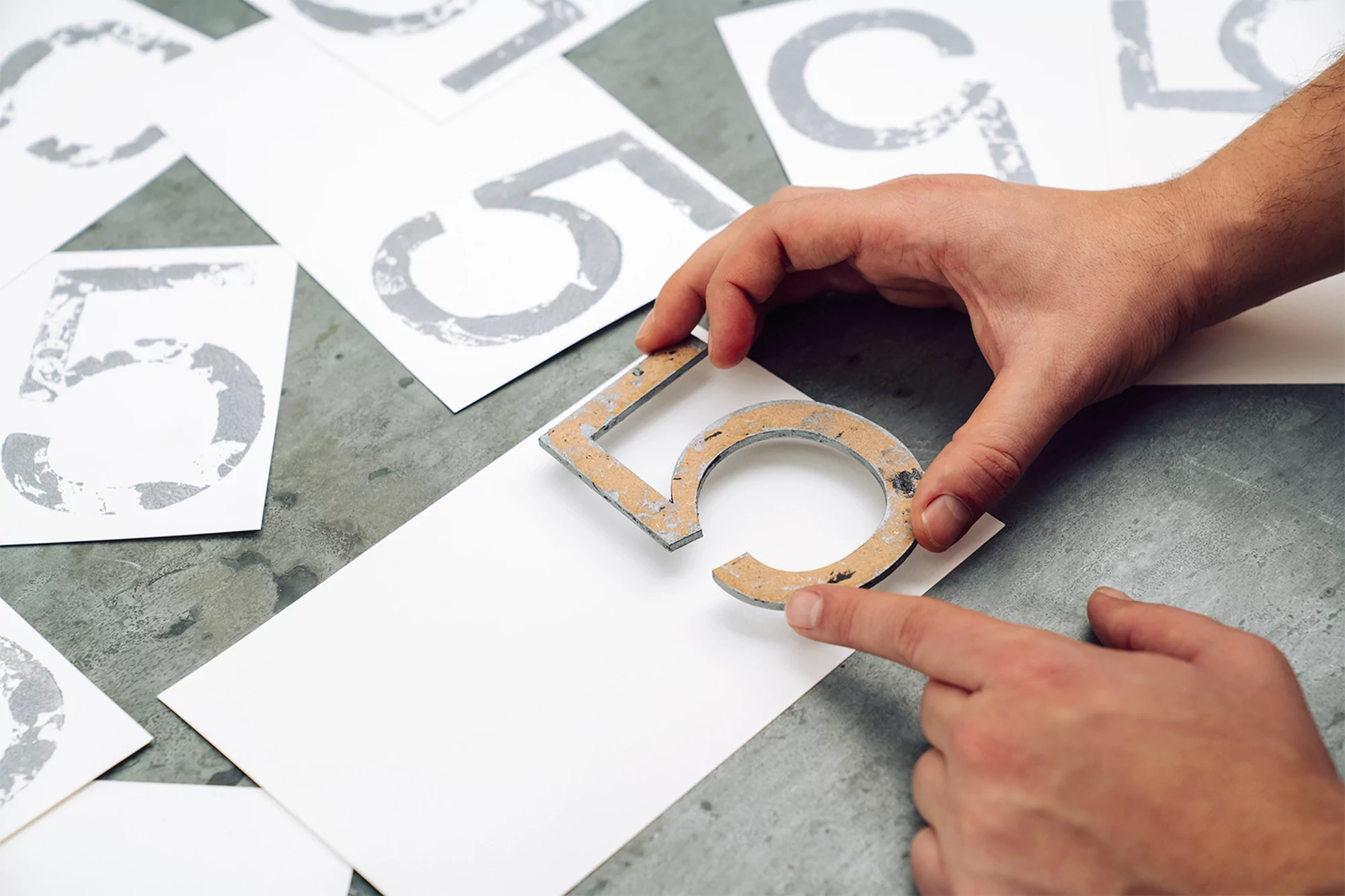 Human hands assemble a number five from layered materials on a blank white card, with more cards printed with gray number fives.