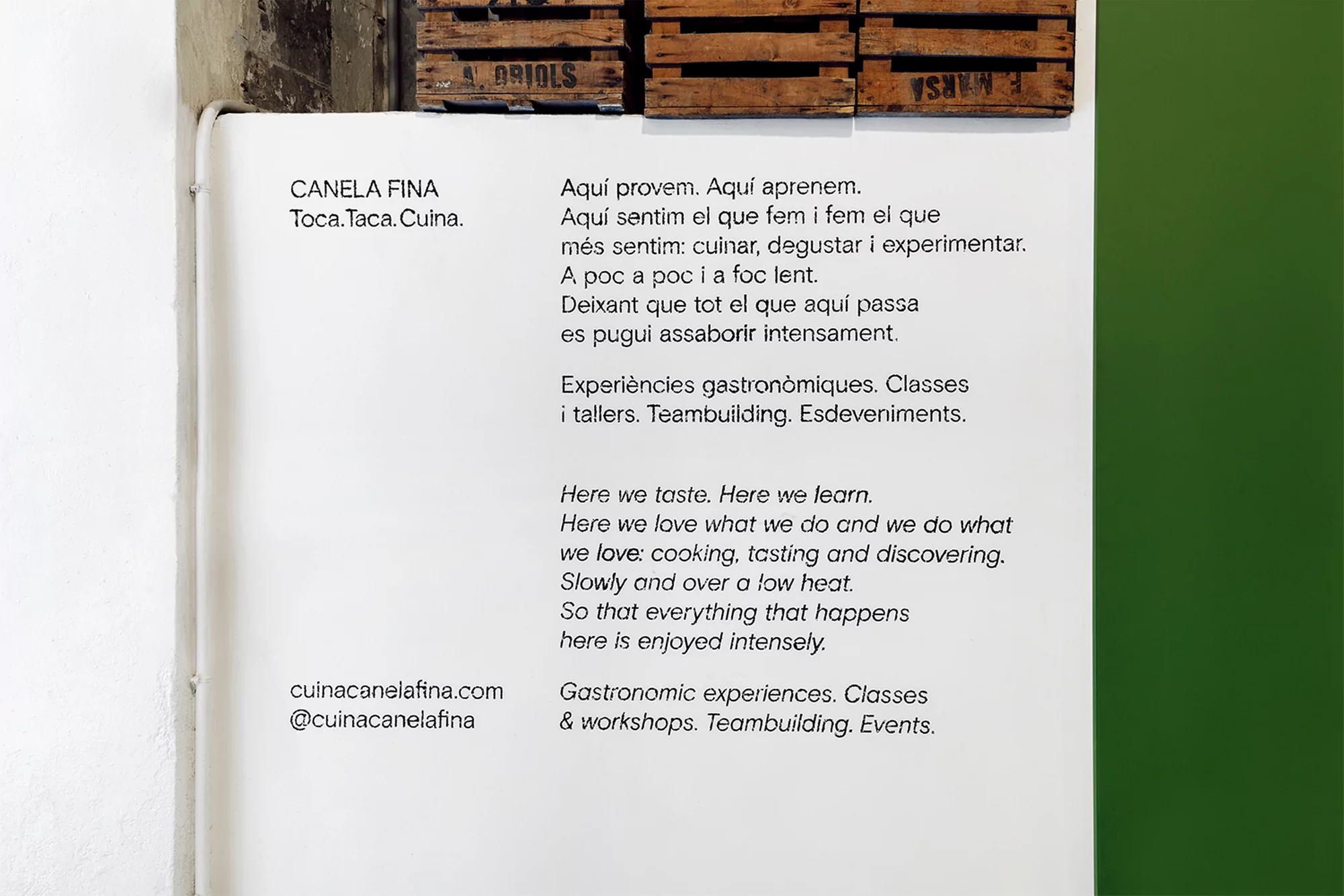 Sign with culinary class descriptions in English and Catalan, featuring the business name and website against a green wall.