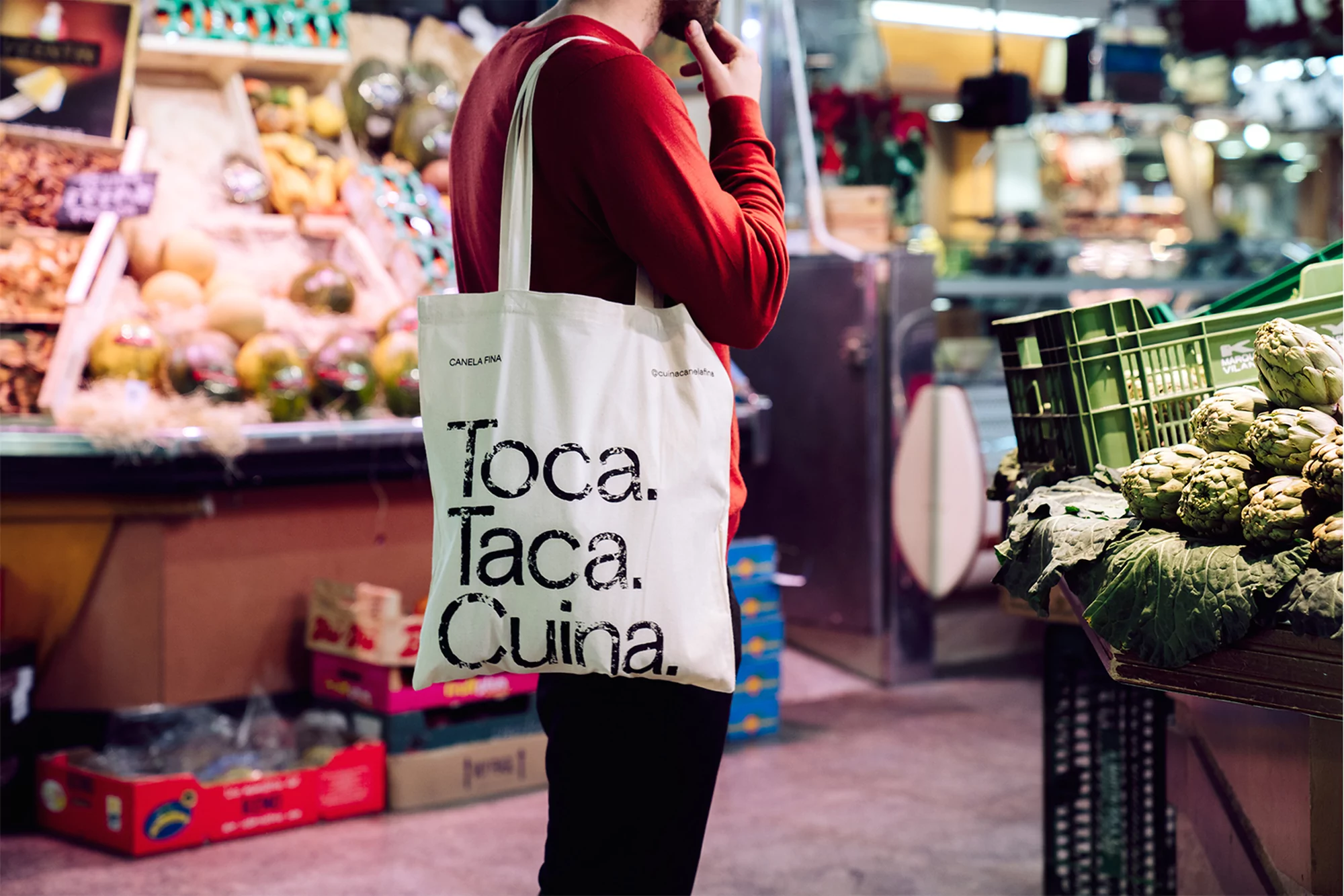 Man in red sweater carries a white tote bag with printed text, standing in a vibrant market with fresh produce and metal shelving.