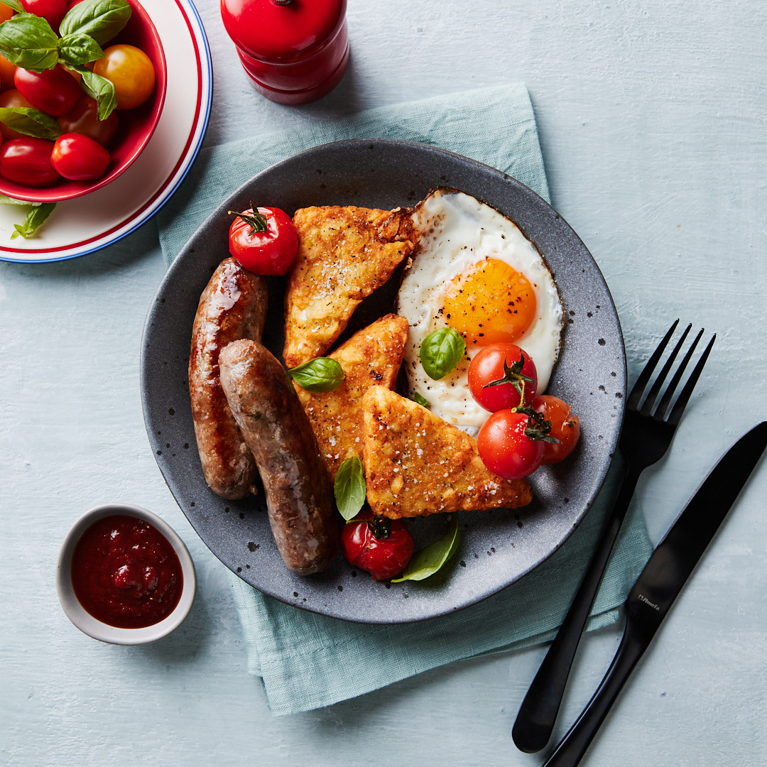 Overhead shot of a breakfast plate with sausages, fried eggs, hash browns, and tomatoes, complemented by sauce, basil, and silverware.