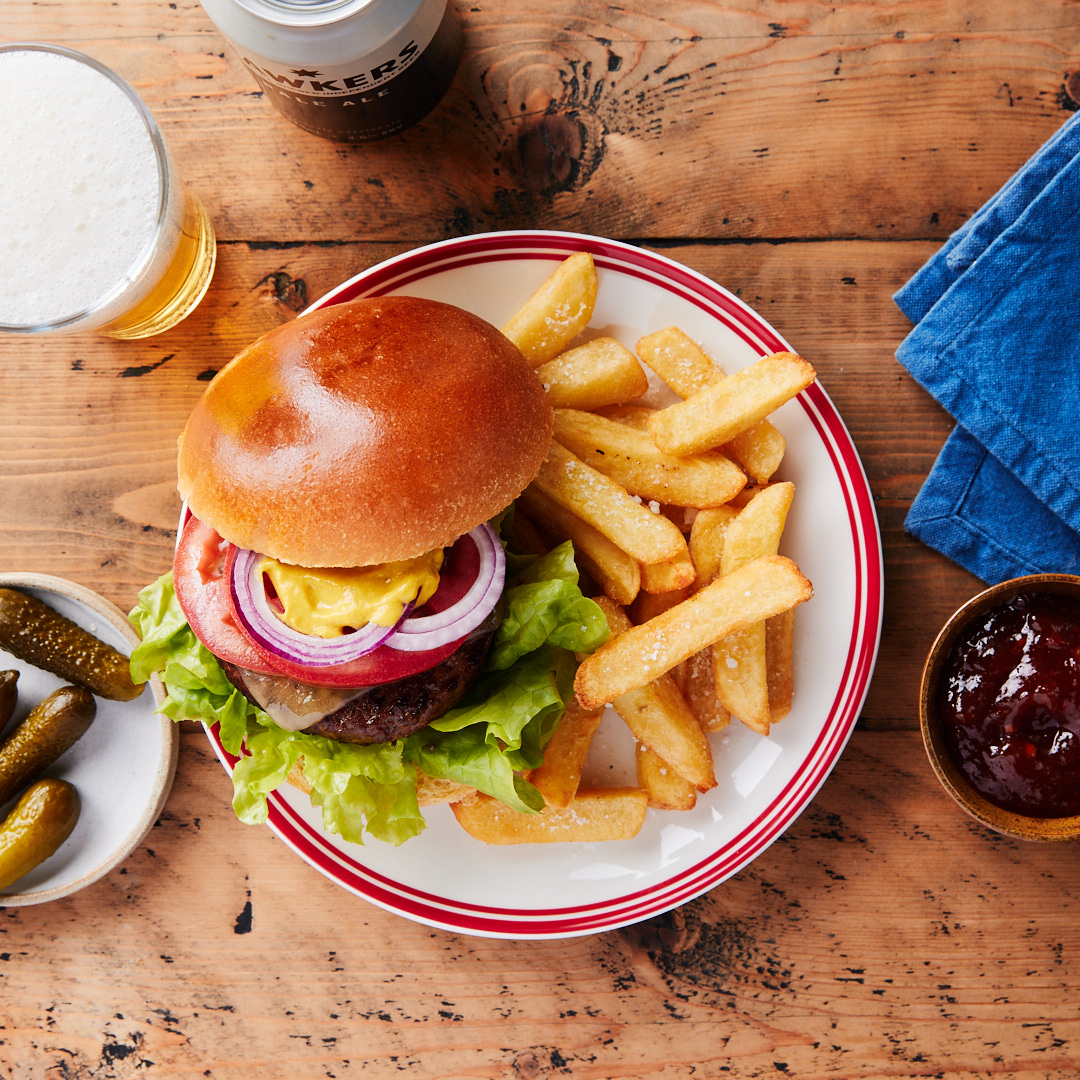 Overhead shot of hamburger with toppings, french fries, beer, pickles, and jam on a wooden table. Food and beverage photography.