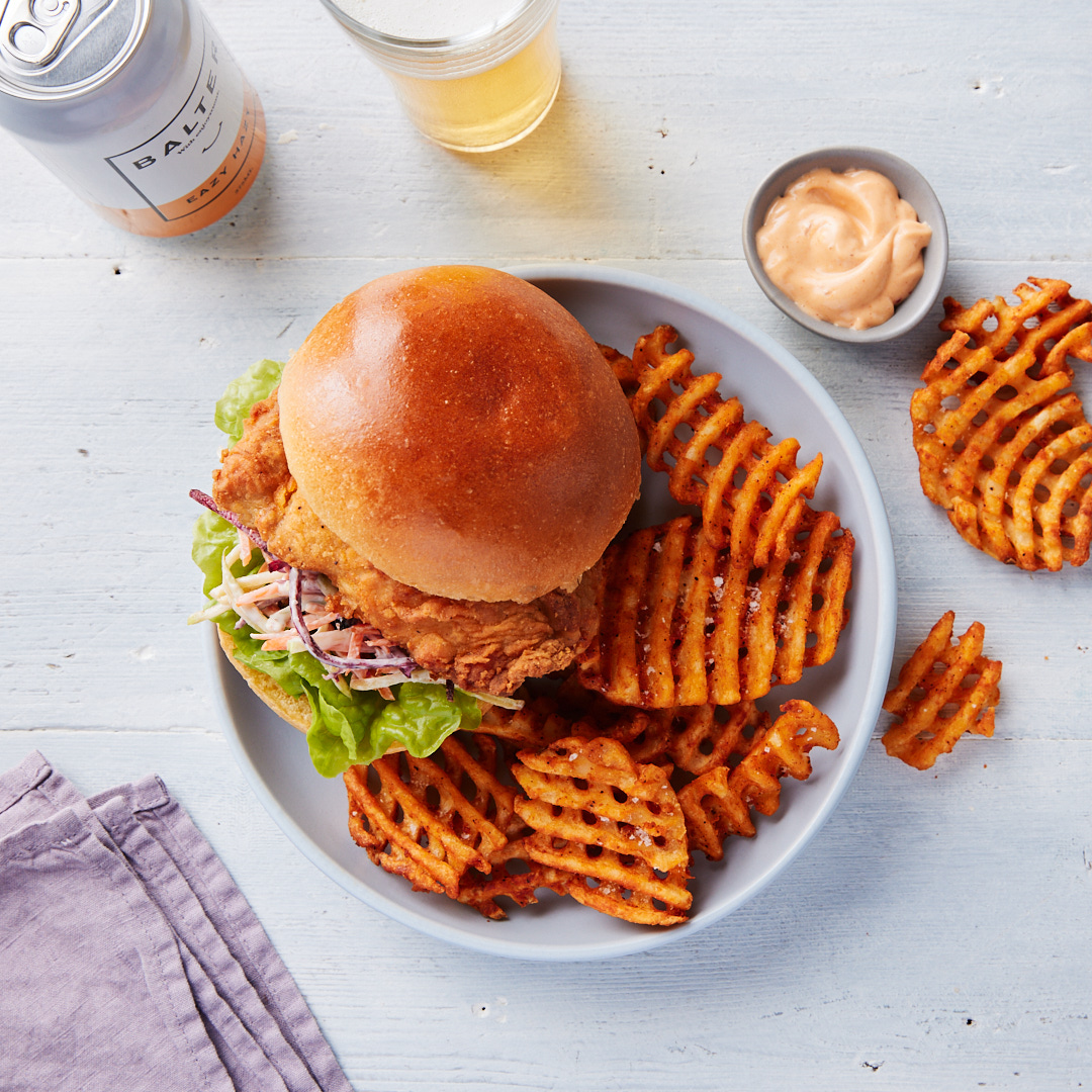 Overhead shot of a chicken sandwich on a bun with waffle fries, dipping sauce, and a can of hard seltzer, staged on a white wood surface.