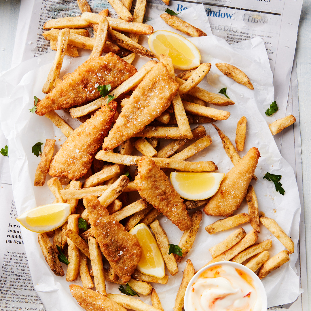 Overhead shot of battered fish and chips with lemon wedges and dipping sauce, presented on white parchment atop a newspaper background.