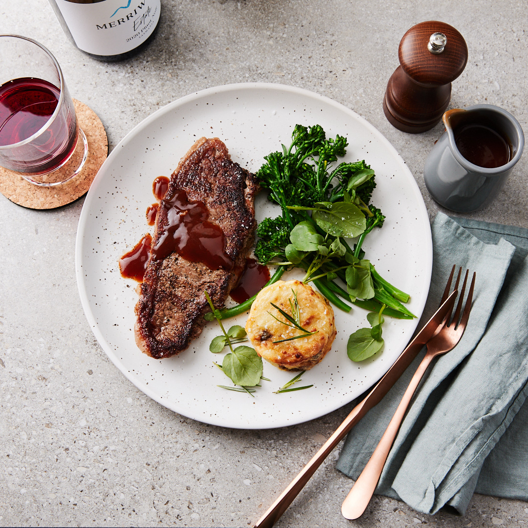 Overhead shot of a steak with red wine sauce and broccoli on a plate, served with potato cake and rosé silverware.