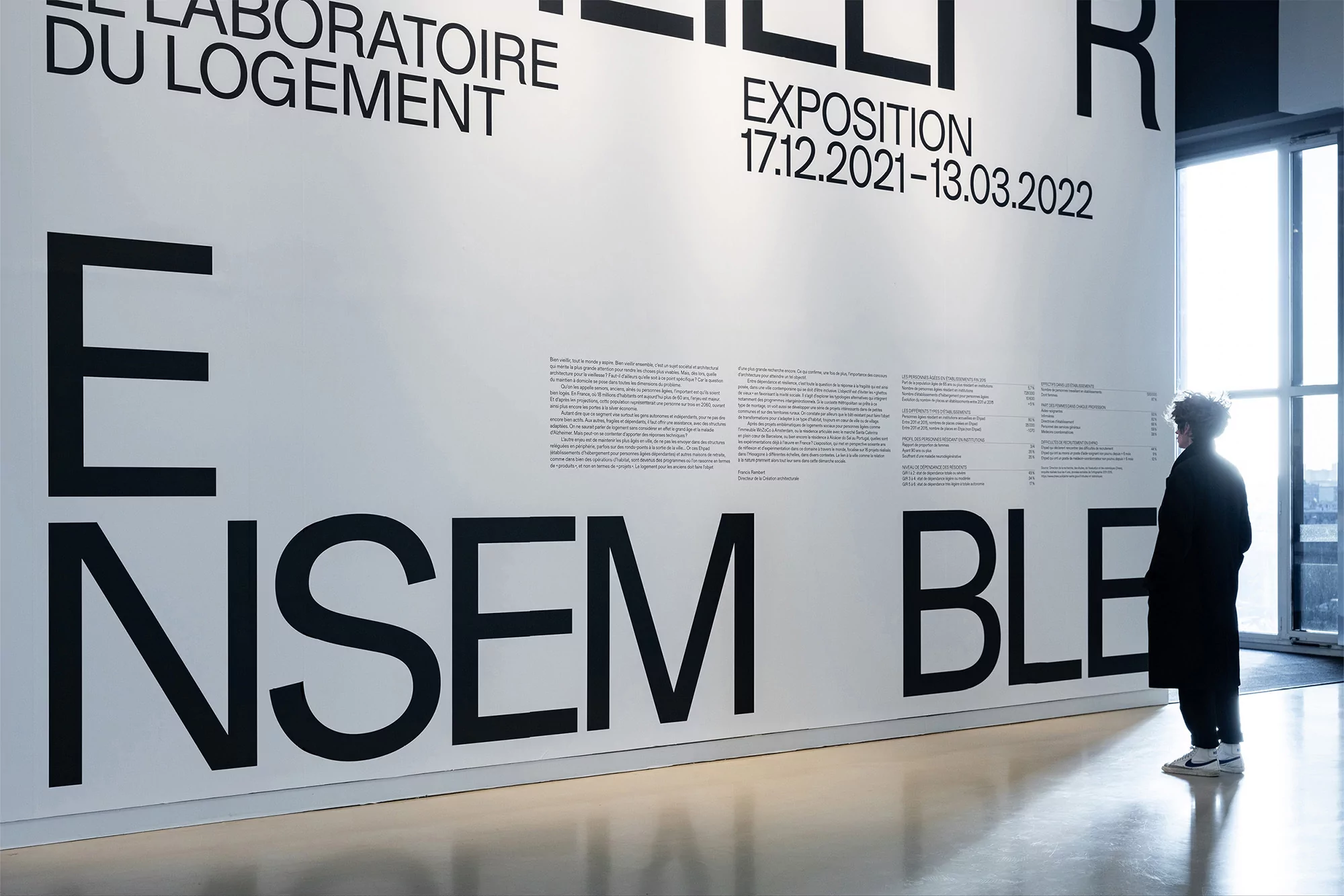 An adult female stands in front of a large poster in a bright room, reading the text on display as natural light streams inside.