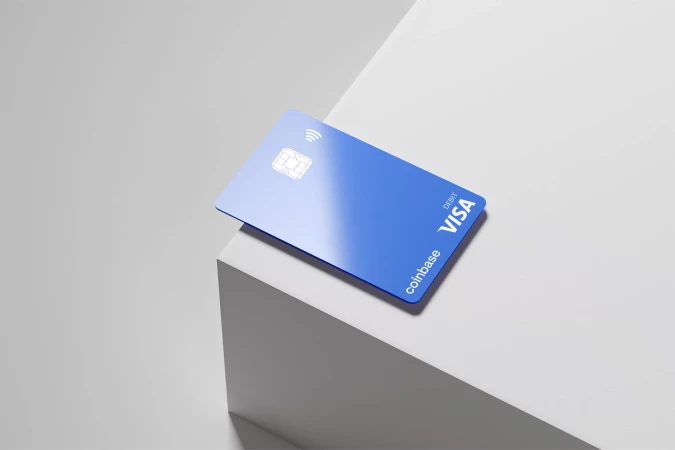 A blue Coinbase debit card on a white geometric platform against a light gray background, showcasing a modern, minimalist financial design.