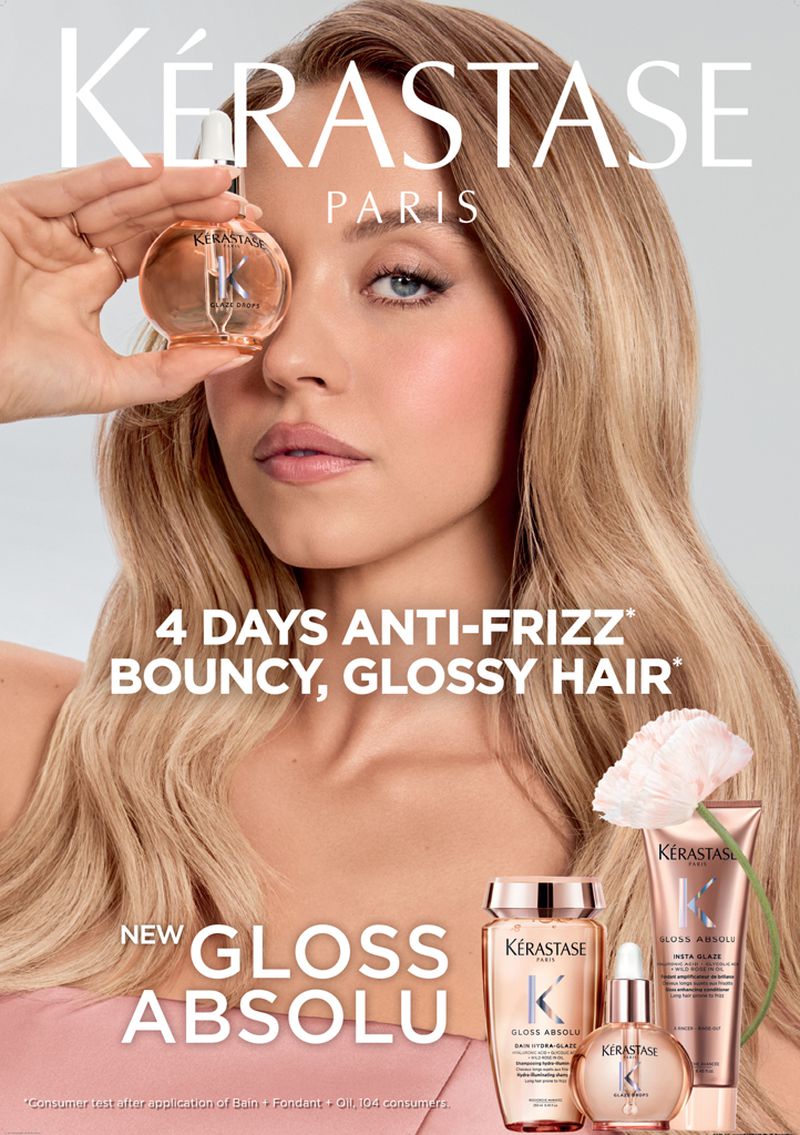 Blonde woman holding hair product bottle with Kerastase brand products, including shampoo and hair oil, in front of light background.