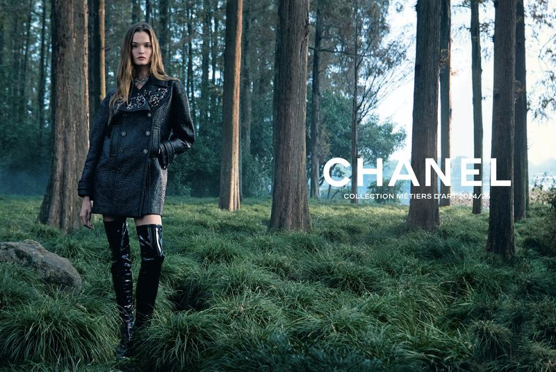 Fashion advertisement in a forest setting featuring a woman wearing a black coat and thigh-high boots, posing near a Chanel logo.