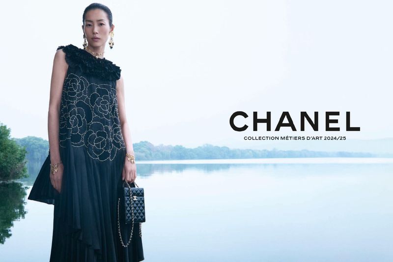 Asian female model in a black floral dress holding a small quilted handbag, posing against a blurred lake background for Chanel ad.