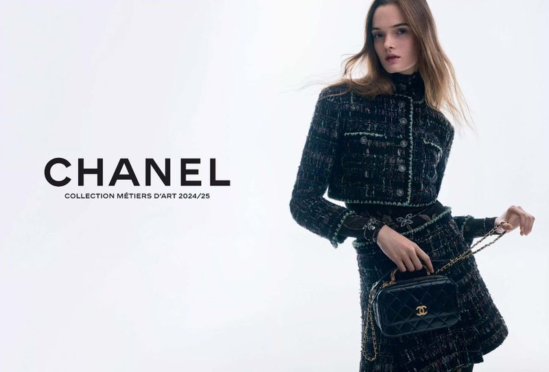 A woman poses in Chanel tweed suit with a black handbag, set against a bright white background, promoting Chanel's Métiers d'Art 2024/25 collection.