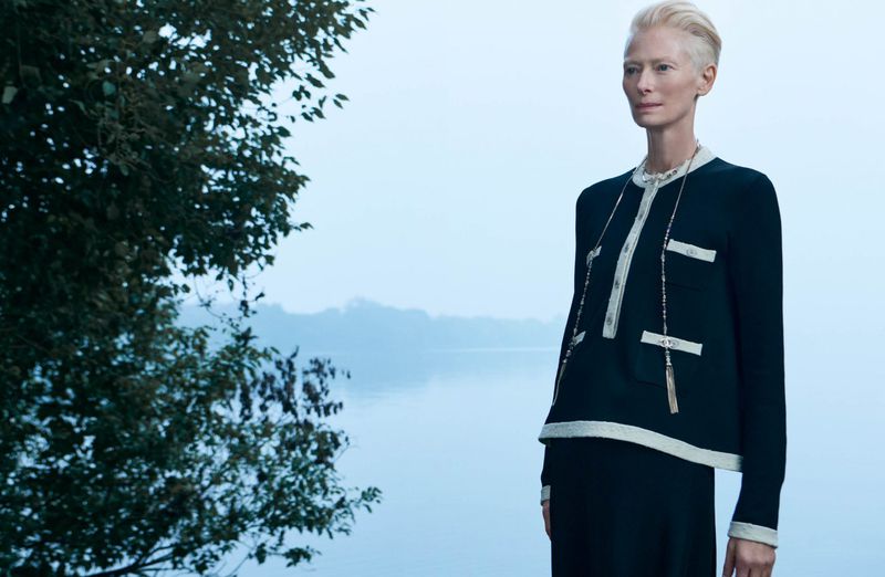 Elegant woman with short blonde hair poses in a black-and-cream jacket and skirt. Lakeside trees create a serene backdrop. 