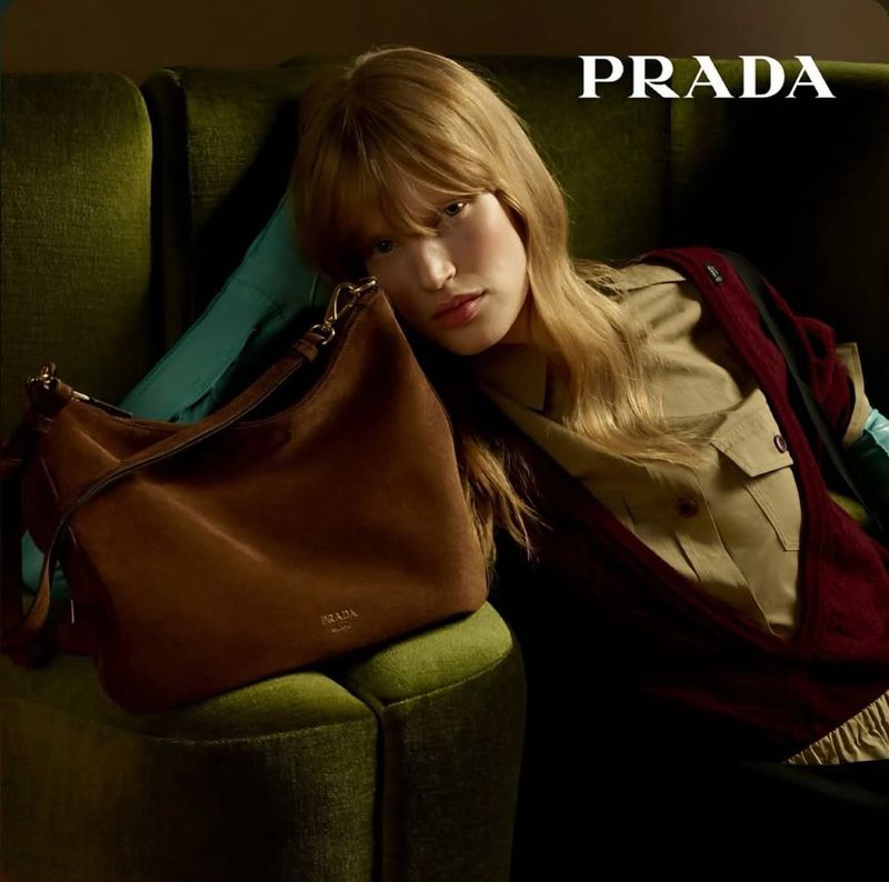 Close-up fashion shot: Woman with blonde hair poses with a brown leather Prada handbag in front of a green velvet sofa.