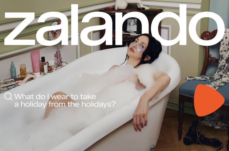 Brunette woman relaxes in a foam-filled bathtub wearing eye mask patches, with beauty products, designer dress and heels in the background.
