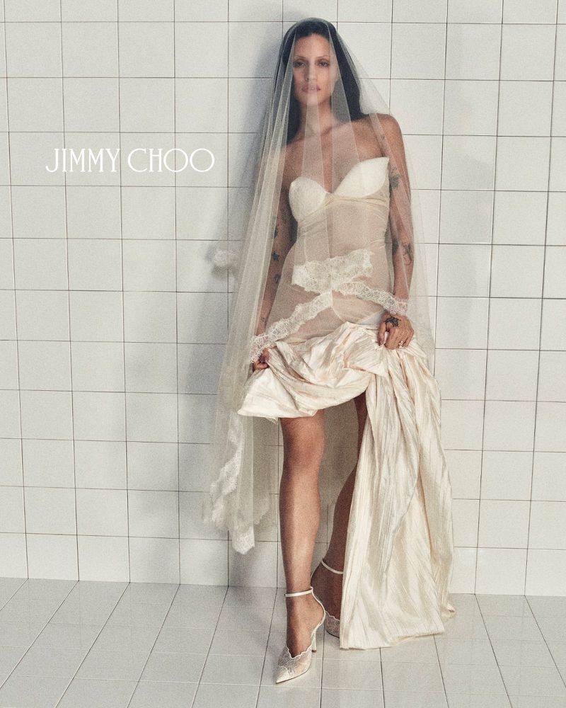 A woman in a wedding dress and veil poses in front of a tiled wall in an indoor environment, showcasing a Jimmy Choo design.