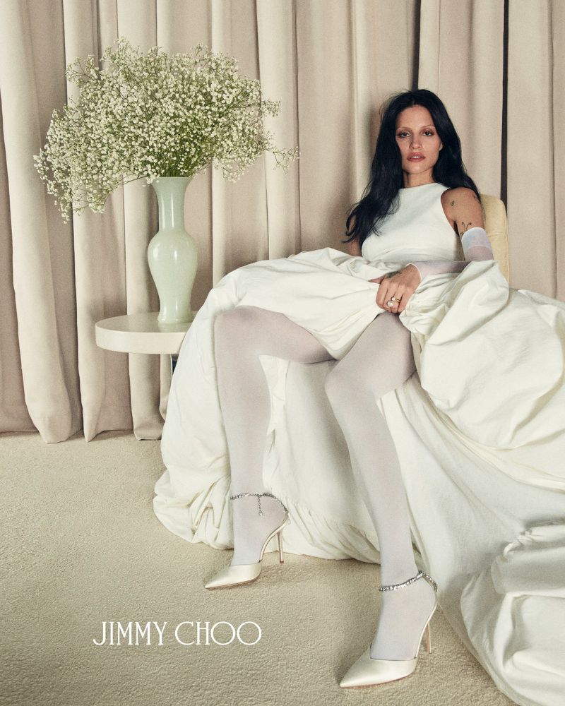 Elegant woman in white dress and Jimmy Choo heels poses with flowers in a minimalist setting, showcasing fashion and style.