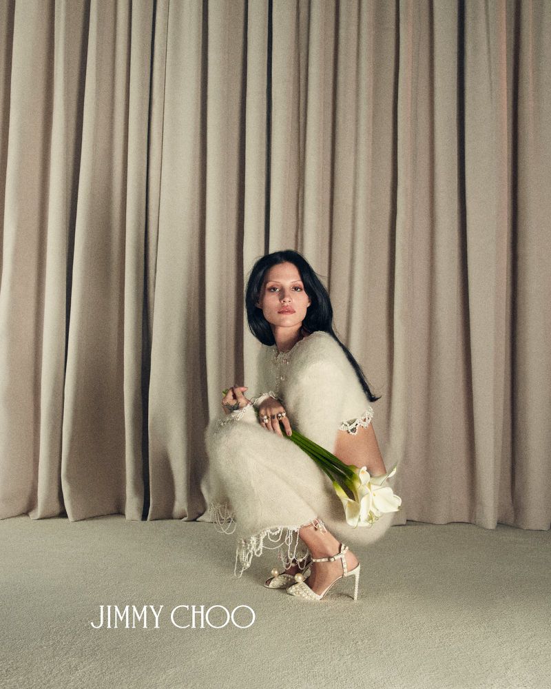 Elegant woman in off-white outfit poses with calla lilies and Jimmy Choo heels against a draped fabric backdrop in a fashion editorial.