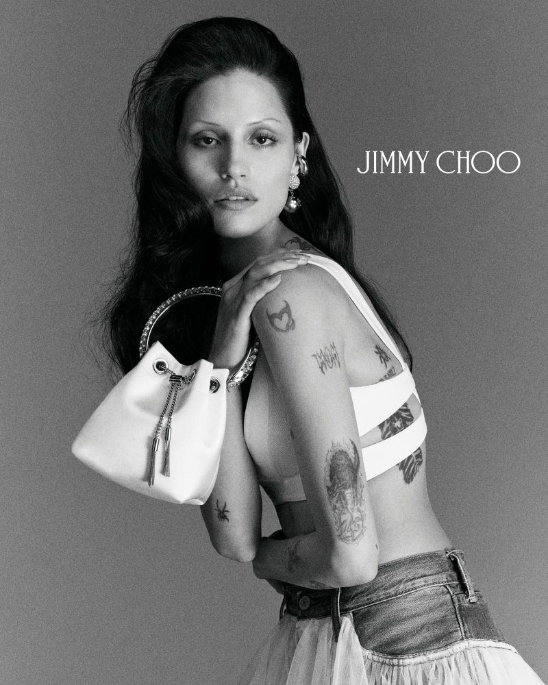 A female fashion model poses with a handbag in a studio, showcasing tattoos and earrings in a black and white Jimmy Choo fashion ad.