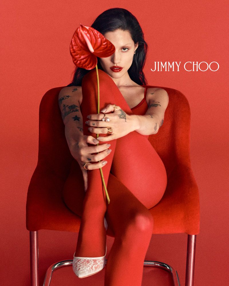 Elegant woman in red dress, tights, and heels sitting on a red chair holding a red flower with Jimmy Choo logo. Fashion-forward.