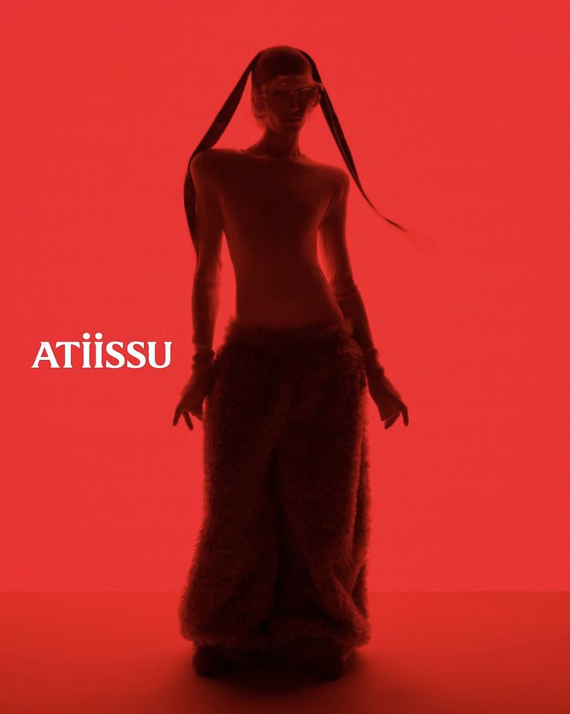 Fashion model in flowing brown outfit stands against a red backdrop in a studio setting. Brand logo 'ATIISSU' visible on left.
