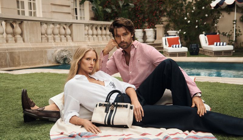 Stylish couple lounging by a pool. The woman wears a white shirt and the man wears a pink striped shirt. Designer bag is in foreground.