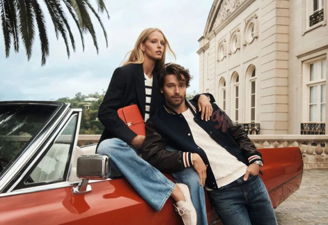 A stylish couple in fashionable attire poses on a red convertible car, set against a backdrop of a building and palm trees.