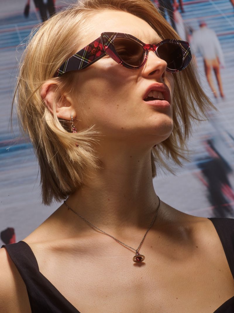 Close-up of an adult female model wearing patterned sunglasses and a necklace, looking upward against a blurred background of people.