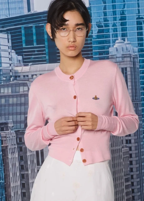 Woman in pink cardigan poses against a city backdrop, showcasing modern fashion and urban style with a soft color palette.