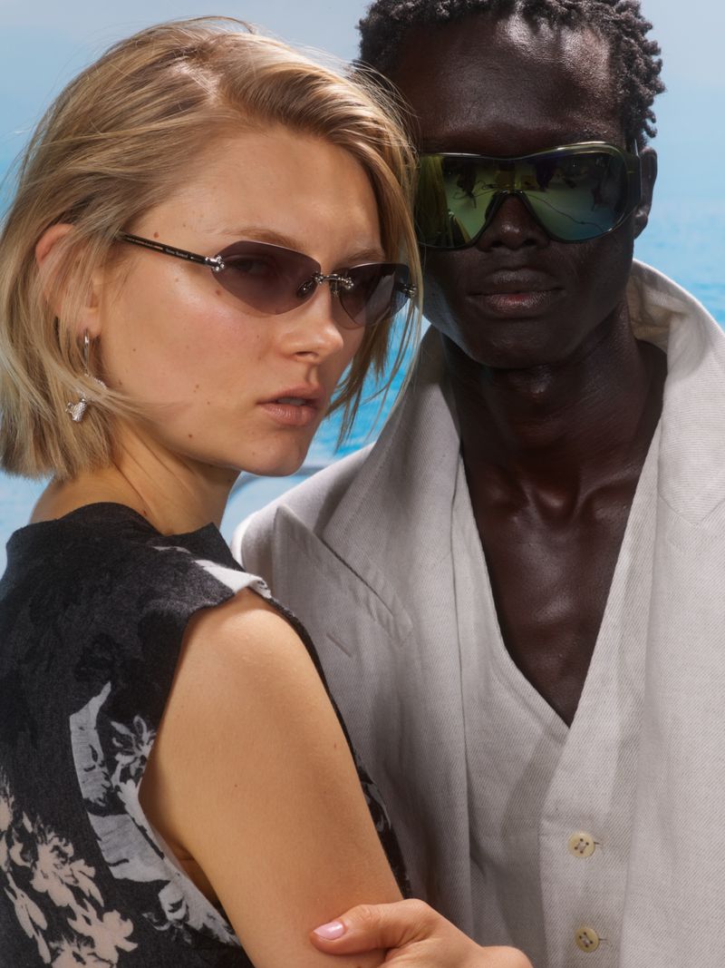 Two fashion models, an African male and a fair-skinned female, pose closely outdoors wearing sunglasses in a commercial shot.