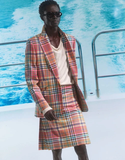 A man poses on a boat wearing a plaid suit with a skirt, sunglasses, and a white t-shirt against a blue water backdrop. Fashion shot.