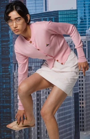 Young adult poses wearing a pink cardigan and cream skirt against a blurred cityscape backdrop. Fashion portrait with modern appeal.
