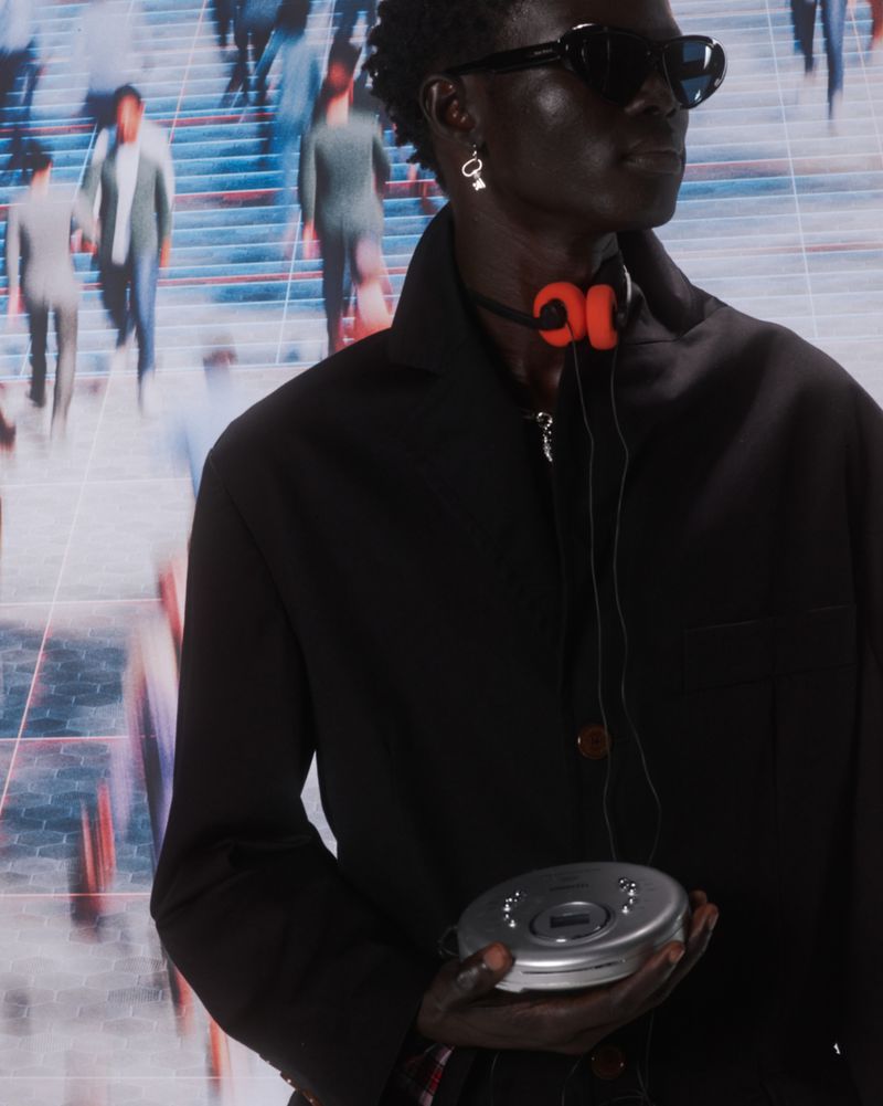 Dark-skinned adult man with an orange headset holds a CD player against a blurry background of people in an urban environment.