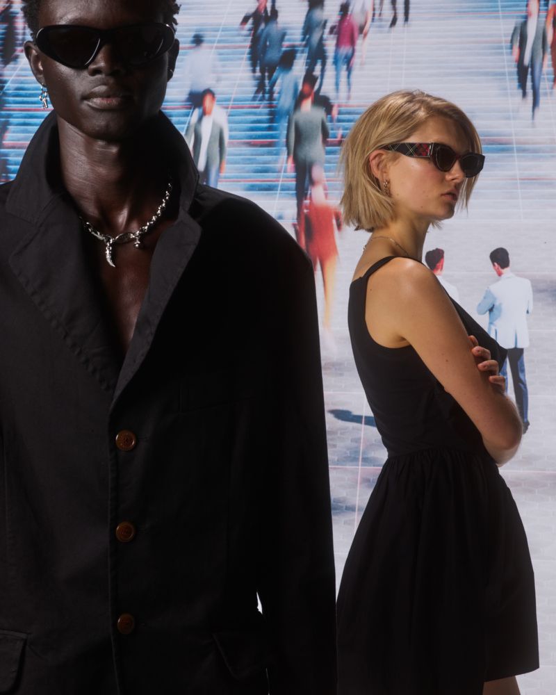 Two fashion models in sunglasses pose against a backdrop featuring blurred figures in an urban scene, shot with studio lighting.