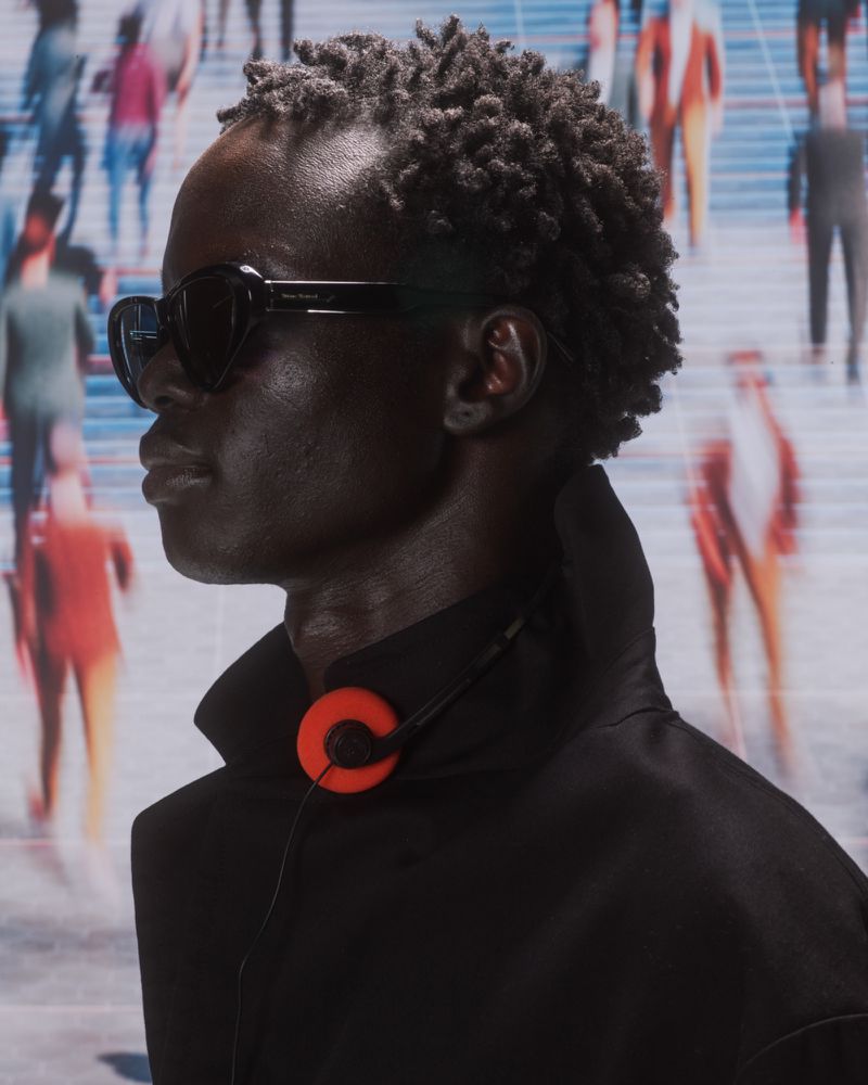 Profile portrait of dark-skinned adult male model wearing sunglasses and headphones, standing against a blurry background of people.